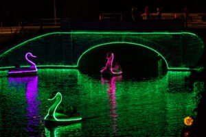 A glowing green bridge over water with illuminated swan and person.