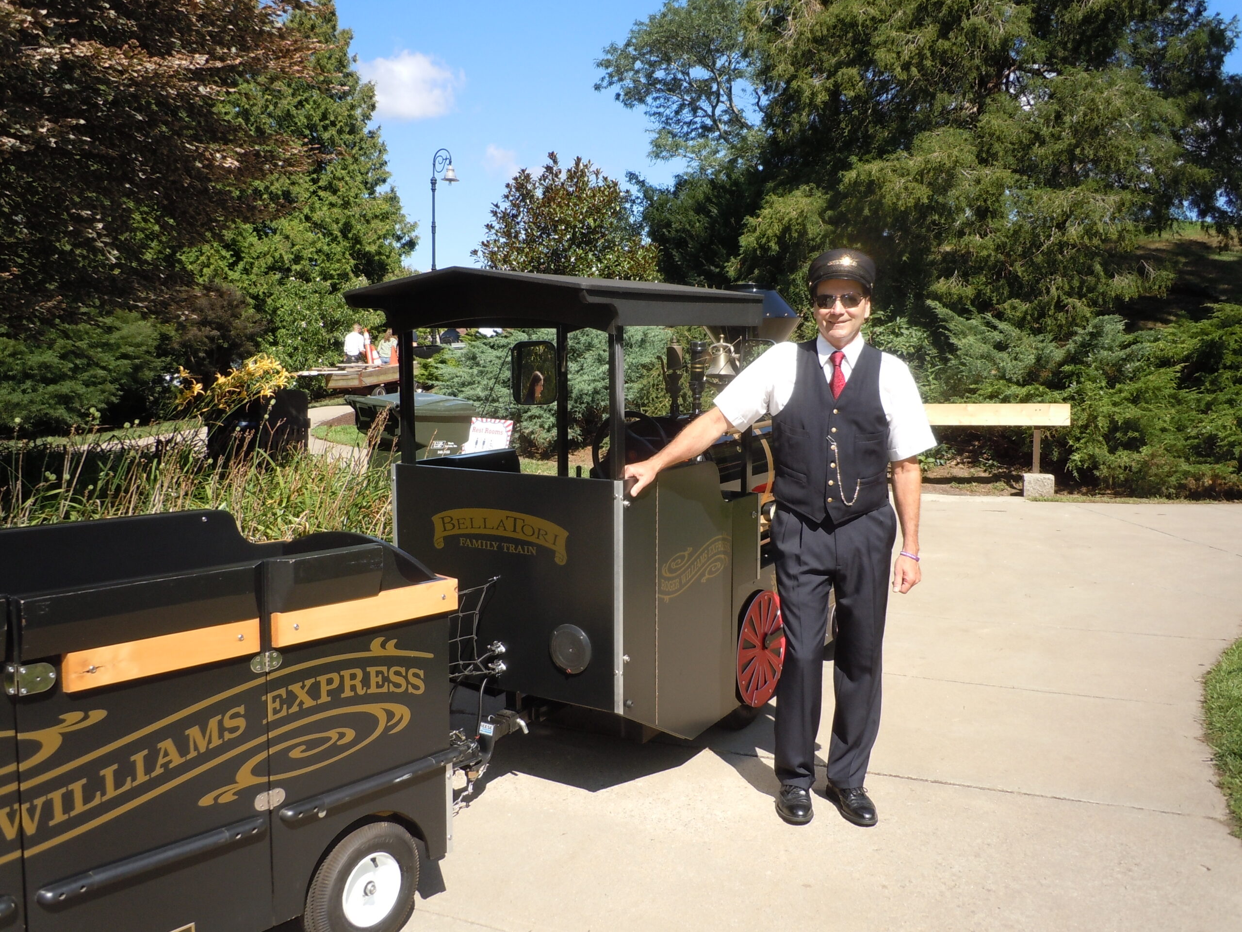 Man in vintage conductor outfit standing by miniature train engine outdoors.
