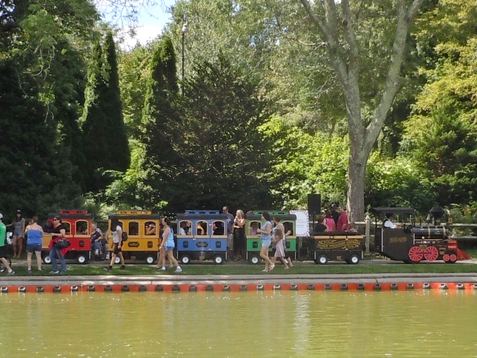 A colorful miniature train rides along a track by a pond in a park.